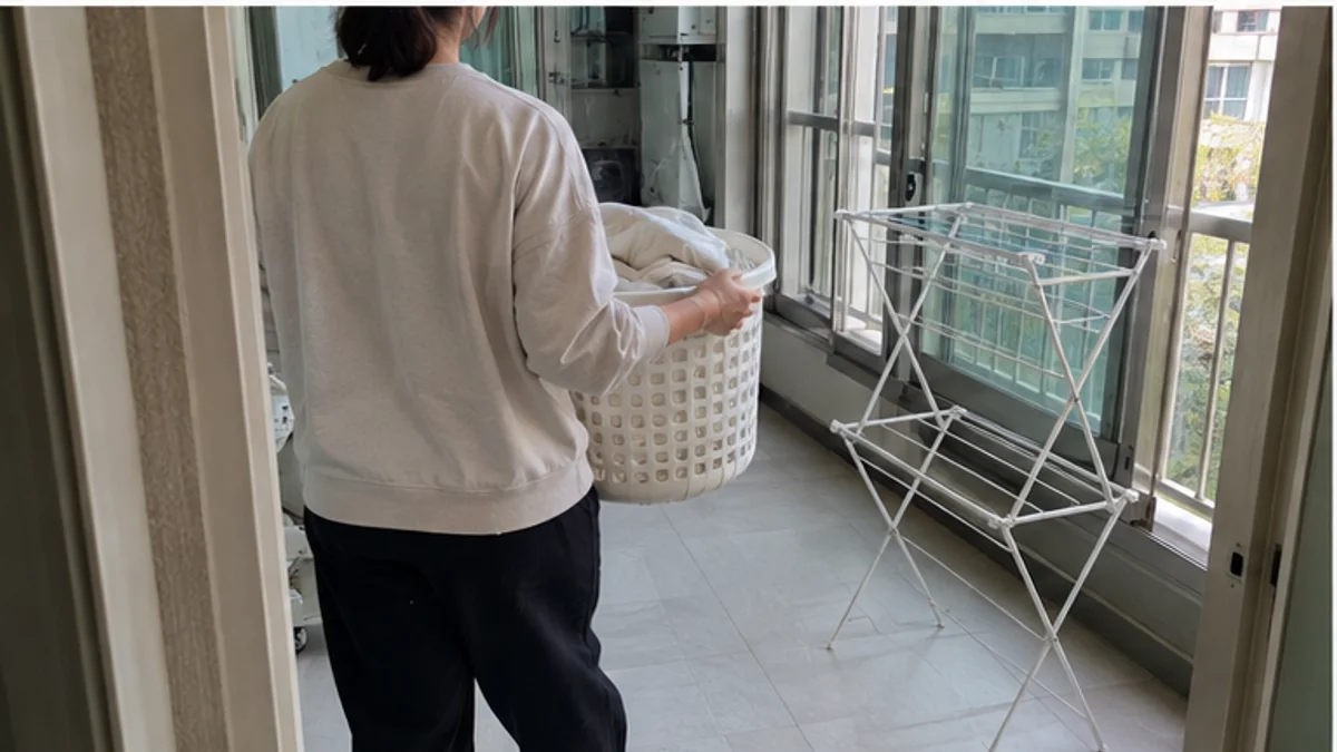 사람이 좁은 베란다 문에서 흰색 세탁 바구니를 들고 펼친 건조대로 걸어가는 모습